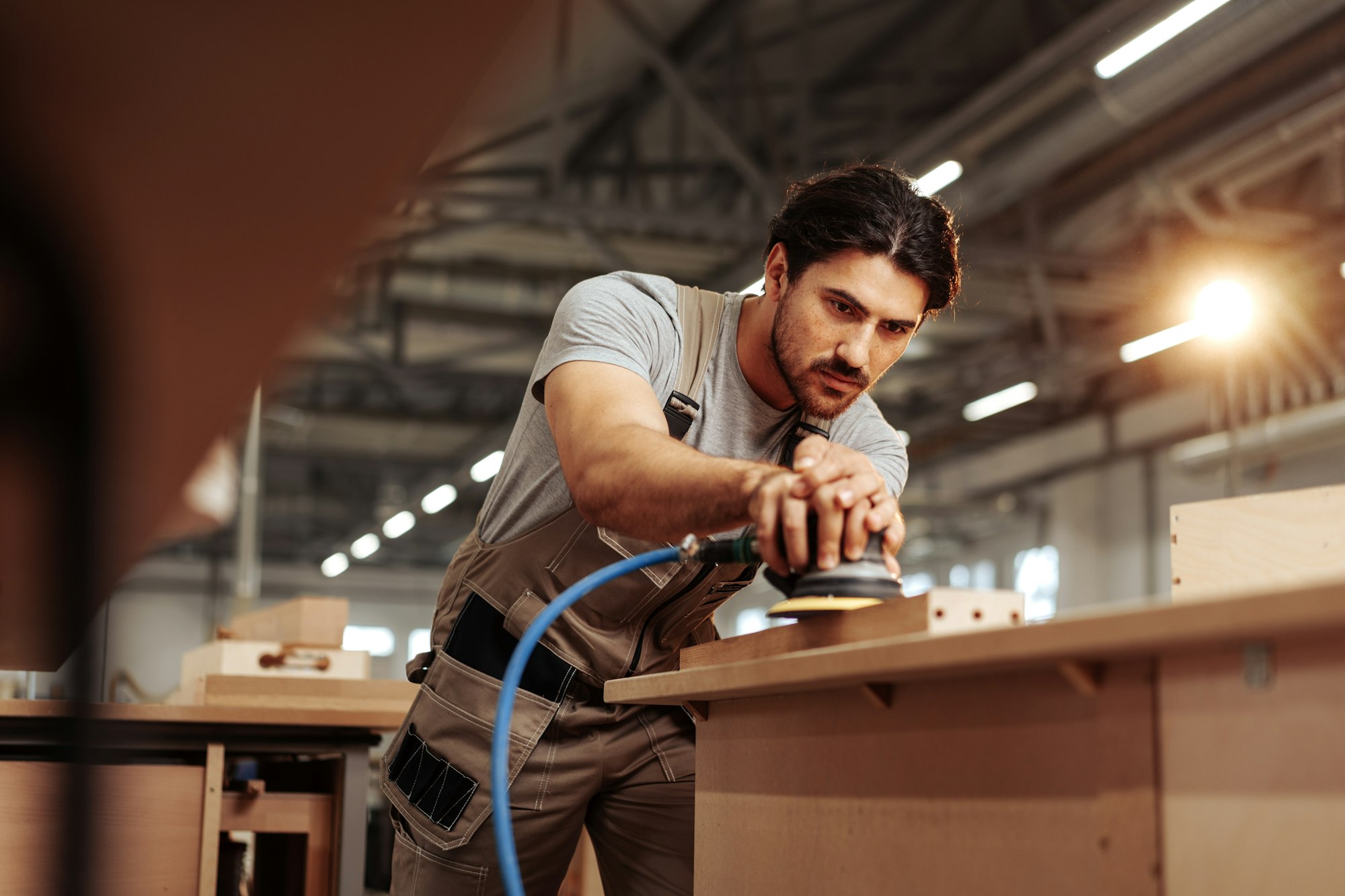 Young carpenter sanding wood piece in workshop in furniture factory